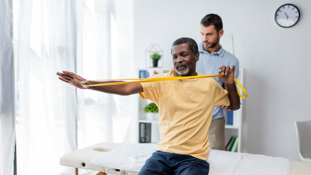 African American man using a rubber exercise band while physical therapist helps.
