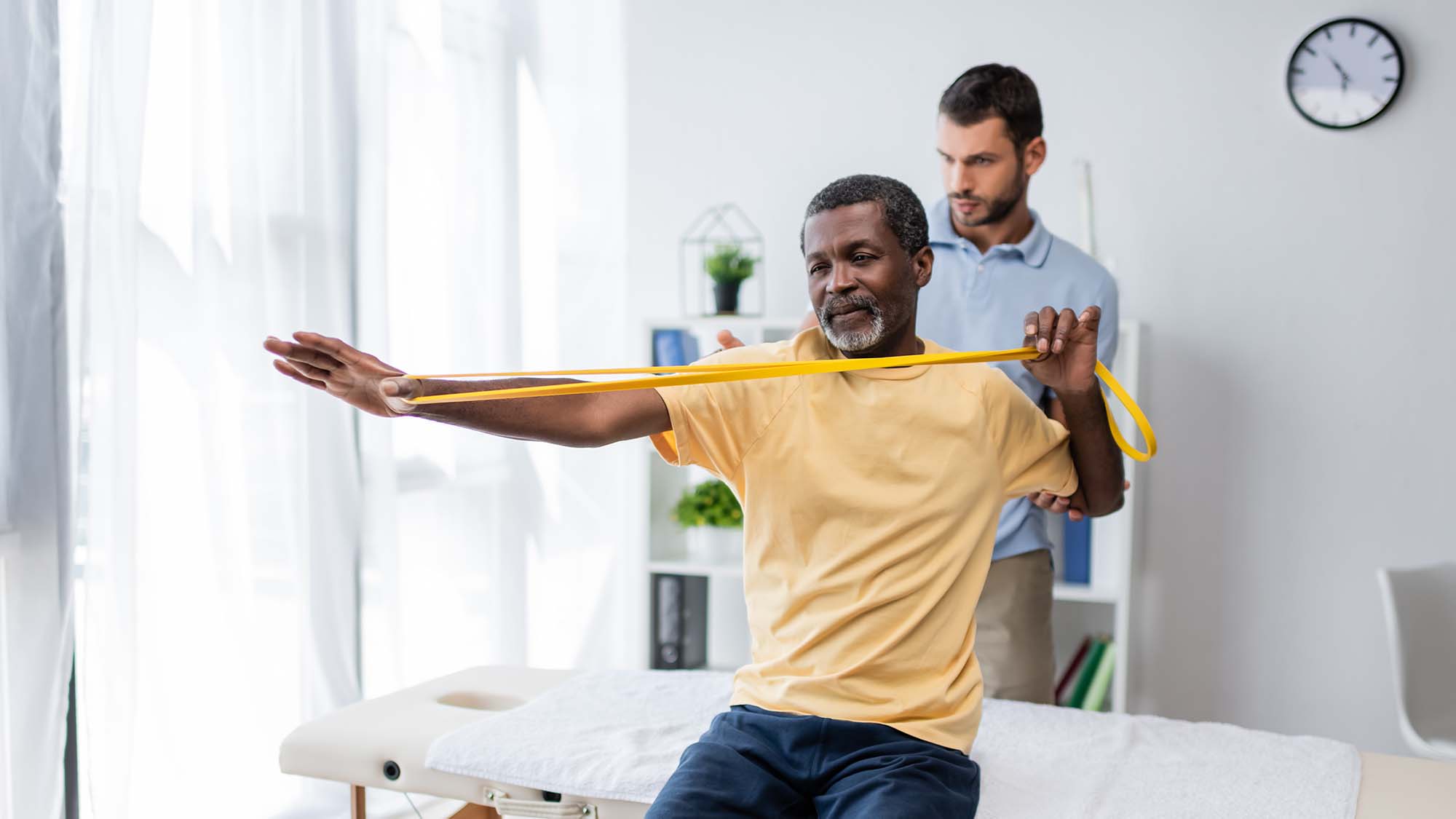 African American man using a rubber exercise band while physical therapist helps.